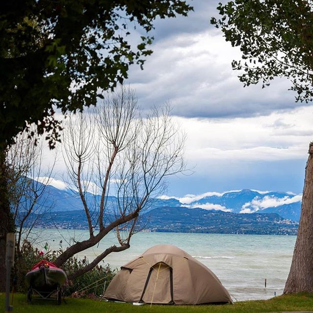 A tent and kayak sit on the green grass near Lake Garda in Italy, with mountains and cloudy skies in the background.