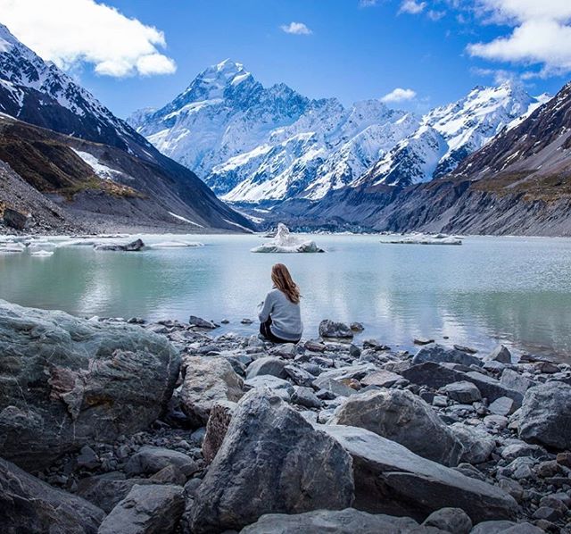 A woman sits on a rocky shore enjoying a mountain lake view on a sunny day.