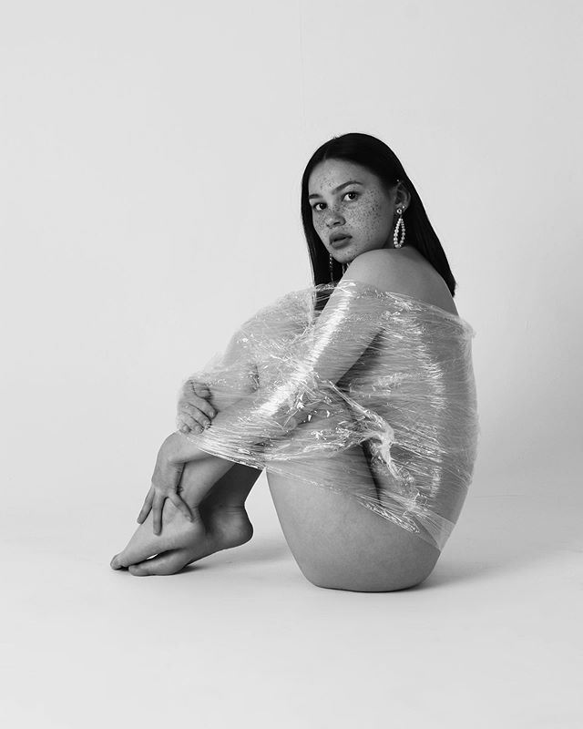 A thoughtful woman wrapped in plastic poses in a minimalist black and white studio shot.