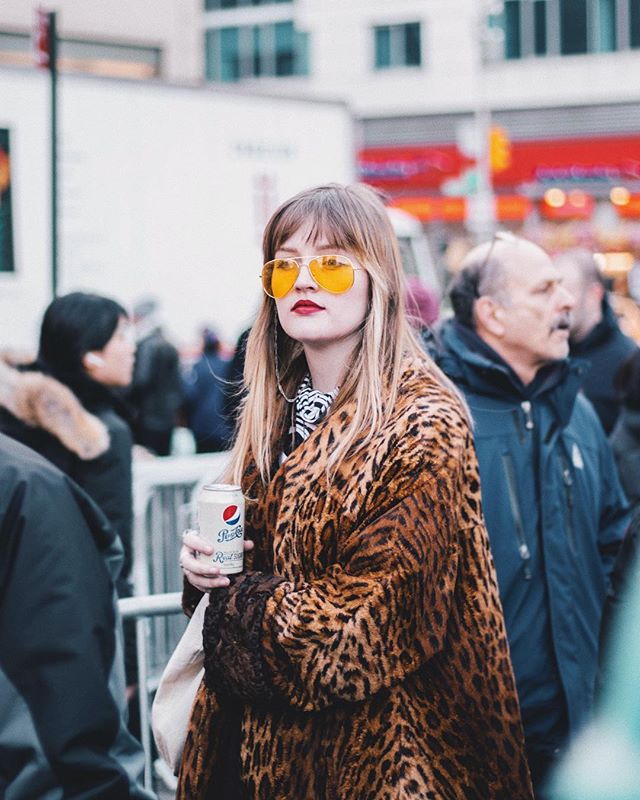 A stylish woman with yellow sunglasses holds a Pepsi can on a busy city street, wearing a leopard print coat.