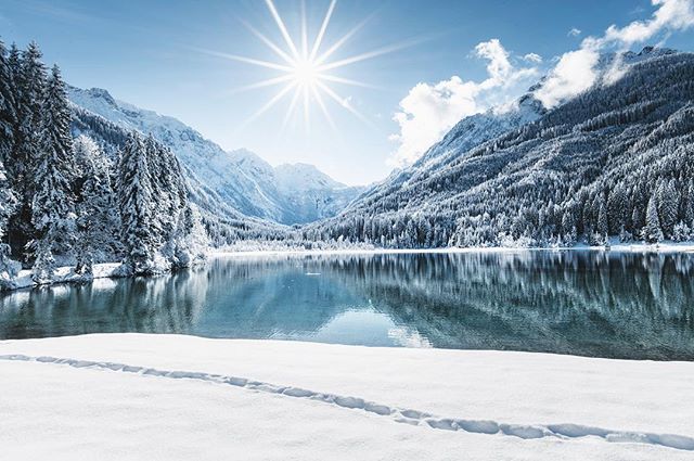 Scenic winter landscape featuring snow-capped mountains reflected in a tranquil lake under a bright, sunny sky.