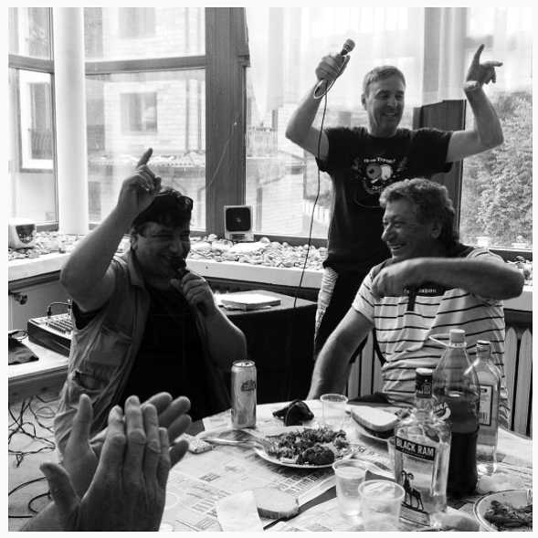 Three men are singing karaoke and celebrating in an indoor setting with drinks and food on the table.