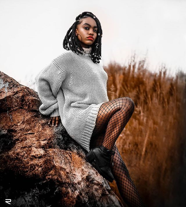 A woman with braids wearing a sweater dress and fishnets poses confidently on a rocky terrain in a fashion editorial.