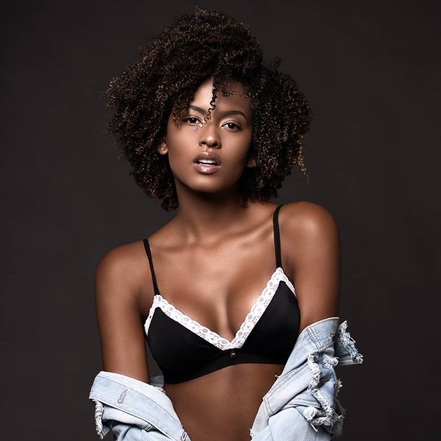A serene African American woman poses in lingerie against a neutral backdrop for a fashion campaign.