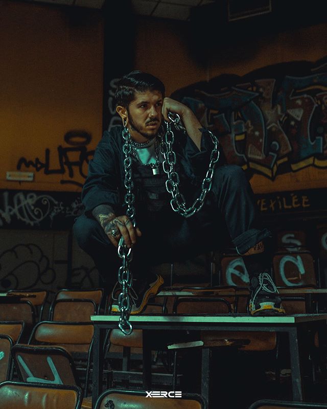 A stylish man with chains sits on desks in an urban classroom setting. He is wearing tattoos, earring and jewlery.