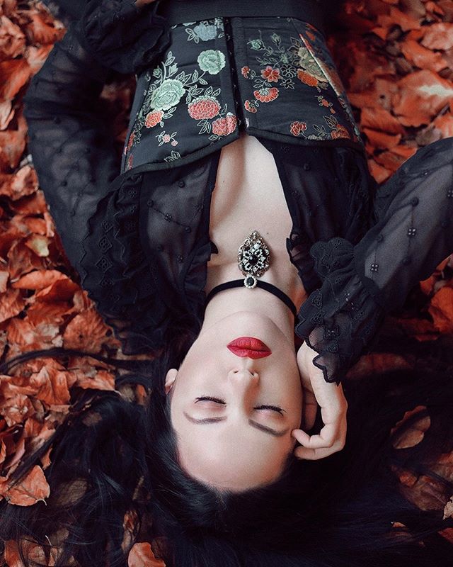 A woman with red lipstick and dark hair lies among fallen leaves in an elegant, moody portrait.
