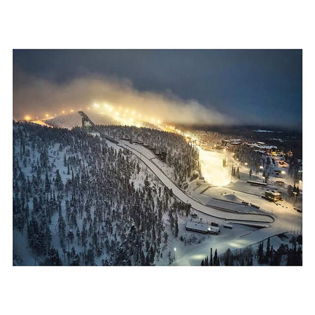 Night view of a snowy ski jump and landscape with trees and lights creating a serene winter scene.