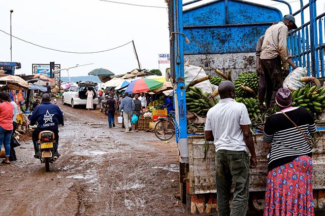 A bustling African market with vendors unloading produce and locals shopping on a muddy street.