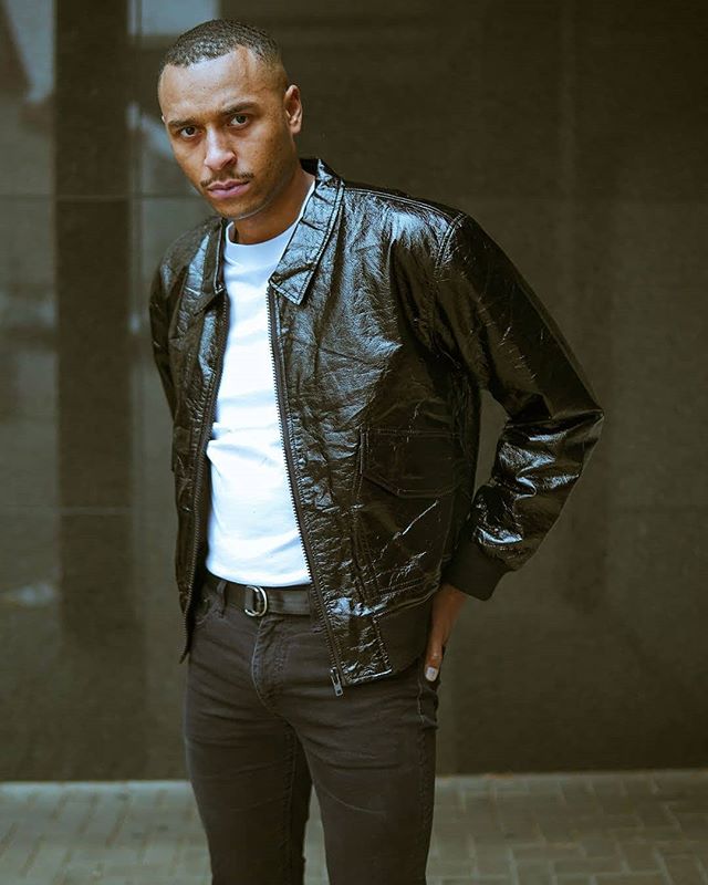 Stylish man in a black jacket and jeans poses against a neutral wall in an urban setting.