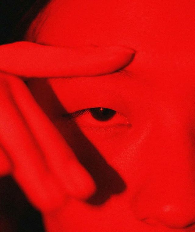 A close-up portrait bathed in vibrant red light focuses on a single eye peering out from behind a hand.
