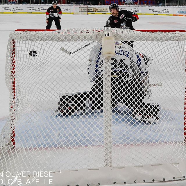 A hockey player and the referee are seen on the ice at a hockey game as the puck flies towards the net.