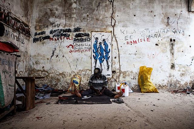 A man sits in a dilapidated room with artwork and writing on the walls, conveying a sense of urban decay and social commentary.