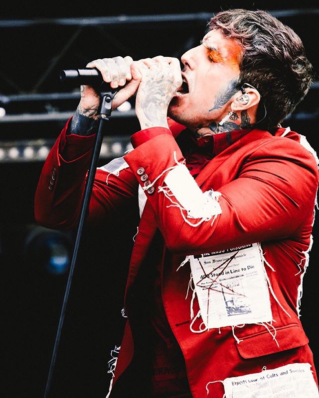 A tattooed singer passionately performs on stage wearing a red suit and singing into a microphone.