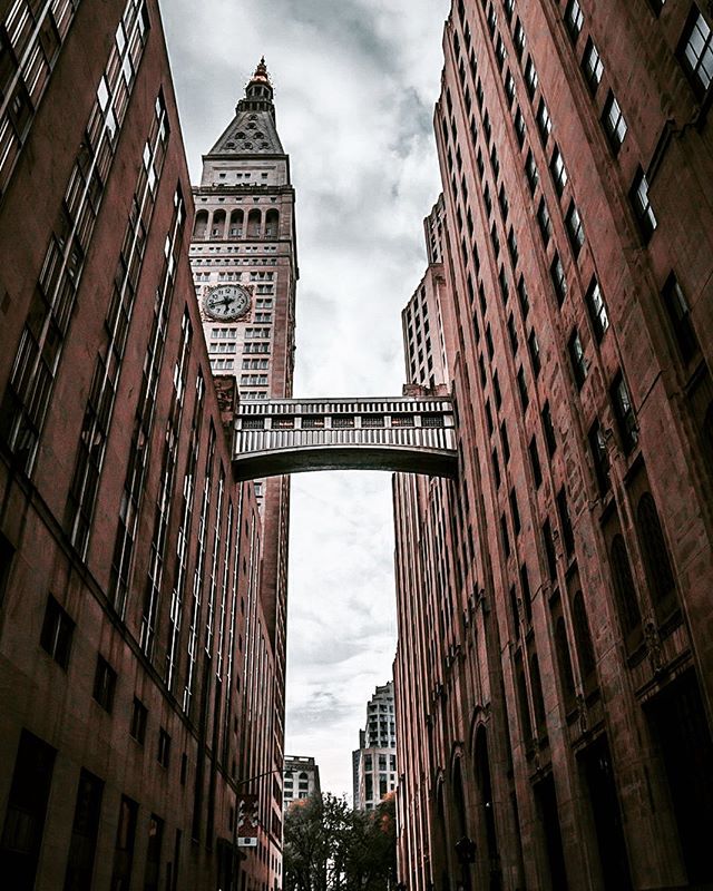 A street view featuring buildings connected by a skybridge with a clock tower in the background.
