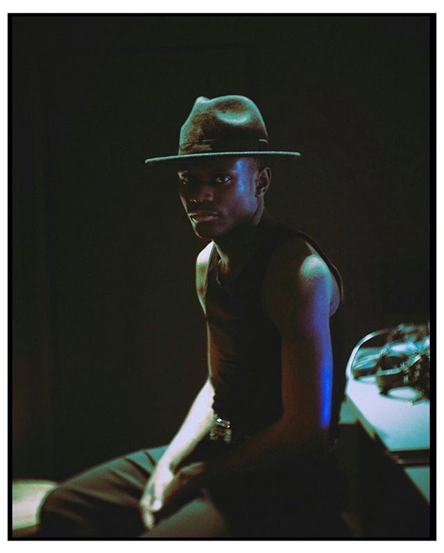 A stylish man in a fedora sits in a studio with cool, moody lighting for a fashion editorial.