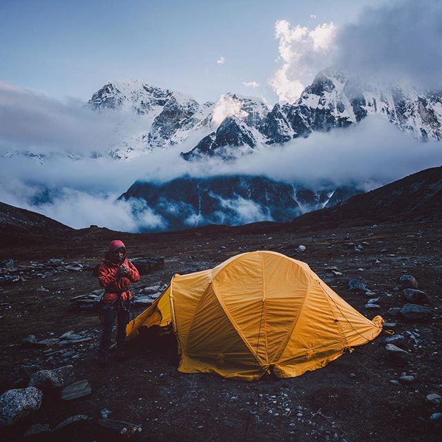 Person stands next to a yellow tent in a rocky landscape with snow-covered mountains and clouds in the background.