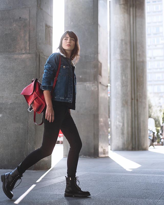 A woman walks confidently in black pants, denim jacket, and a red backpack on a city street.