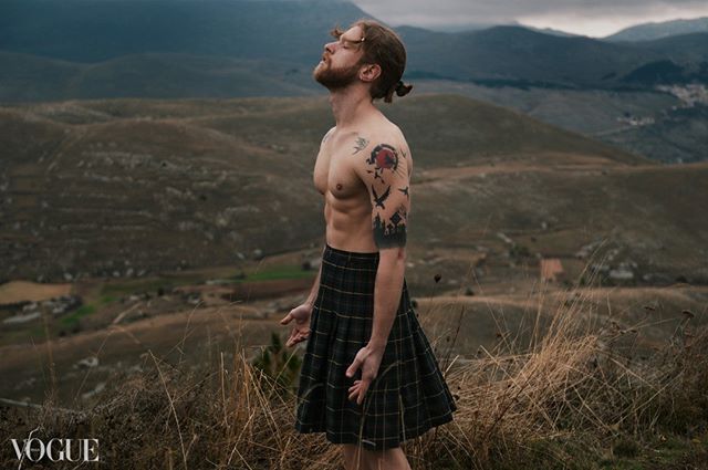 A man with tattoos wearing a kilt stands in a mountain landscape.