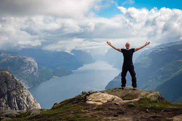 A bald man stands on a cliff with arms raised, overlooking a scenic fjord and mountain landscape.
