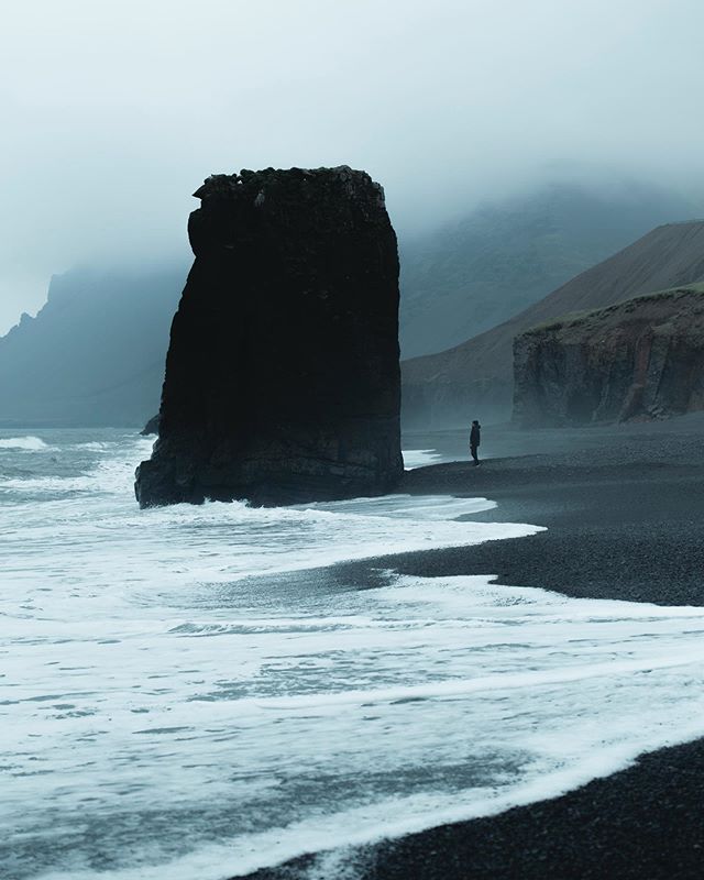 A solitary figure stands on a black sand beach next to a striking rock formation on a misty day.