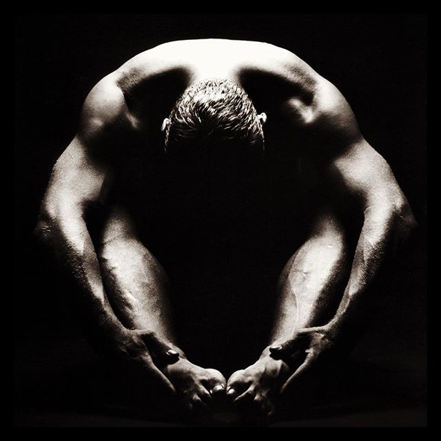 A muscular man sits with his head down, showcasing his physique in a high-contrast monochrome studio shot.
