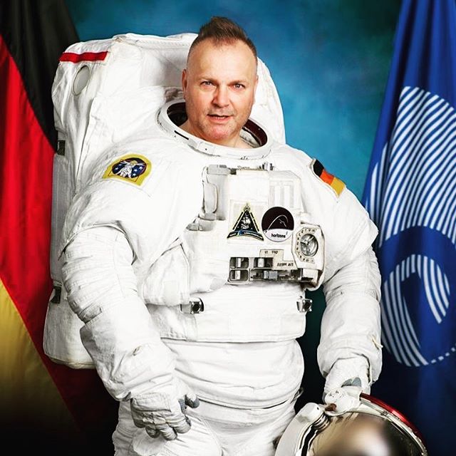 An astronaut poses in a spacesuit next to the German and EU flag. 