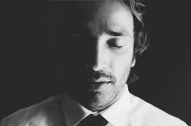 A man with closed eyes in a white shirt and tie, captured in a high-contrast black and white studio portrait.