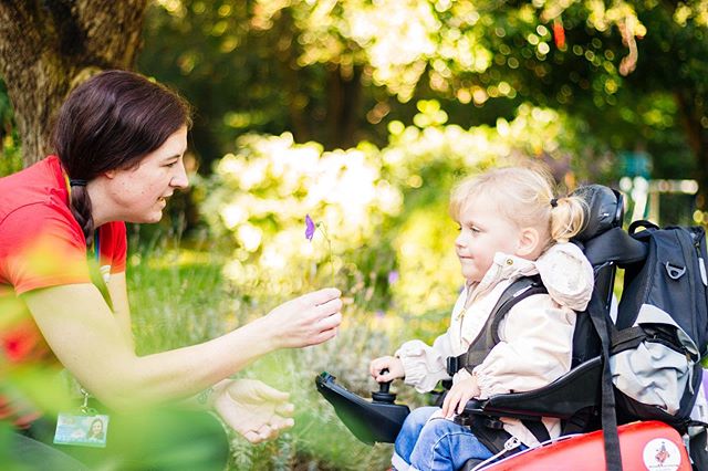 A woman shares a purple flower with a child in a wheelchair, in a sunny park.