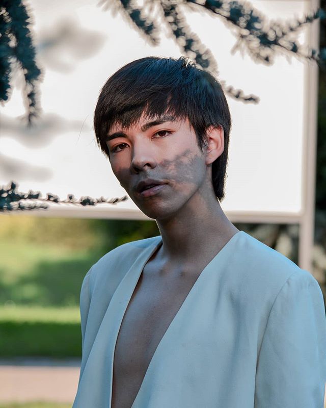 Portrait of a young Asian man in a light jacket with dappled light on his face in a garden setting.