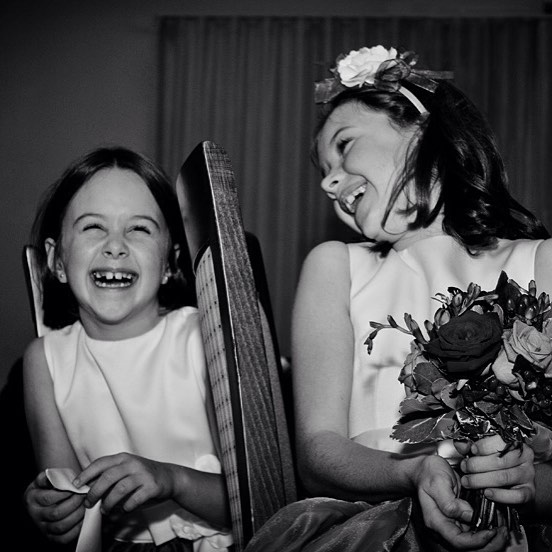 Two flower girls share a joyful moment at a wedding, laughing together in a candid black and white shot.