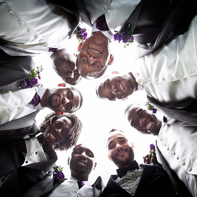 A group of formally dressed groomsmen smiling and looking down at the camera in a low angle at a wedding.