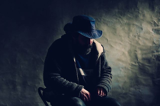 A man with a beard sits pensively in a dimly lit studio, wearing a hat and sweater.