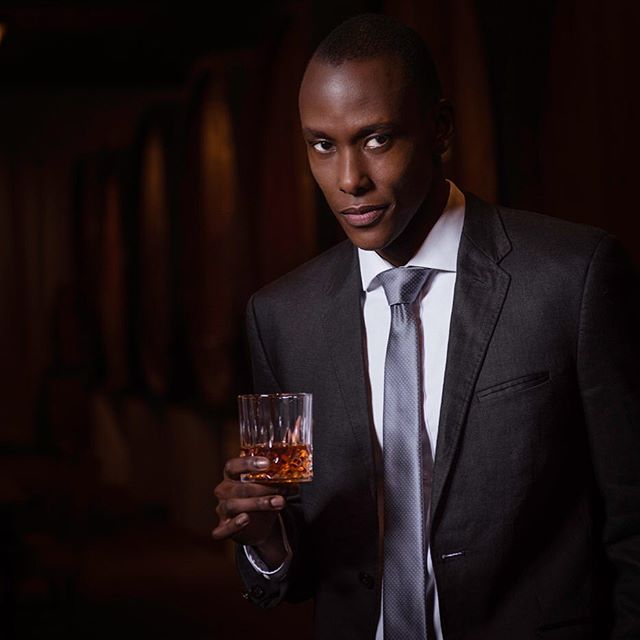 A stylish man in a suit holds a glass of whiskey, exuding confidence and class in a dimly lit setting.