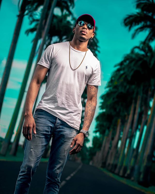 A stylish man poses on a palm-lined road in jeans and a t-shirt, showcasing urban fashion and confidence.