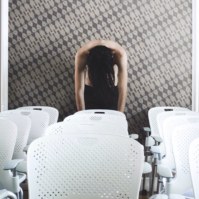 A person bends backward in a room full of white chairs against patterned wallpaper.