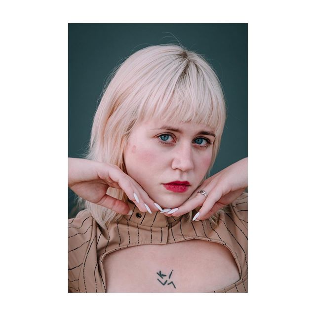 A portrait of a blonde woman with blue eyes and a tattoo on her chest.