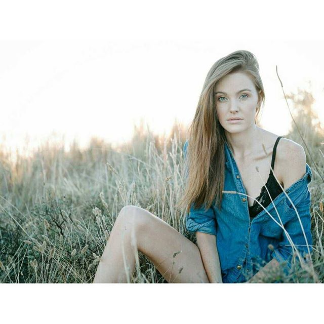 A woman in a denim shirt sits in a grassy field, exuding natural beauty in soft, dreamy light.