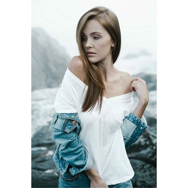 A young woman poses in a white shirt and denim jacket against a rocky mountain backdrop.