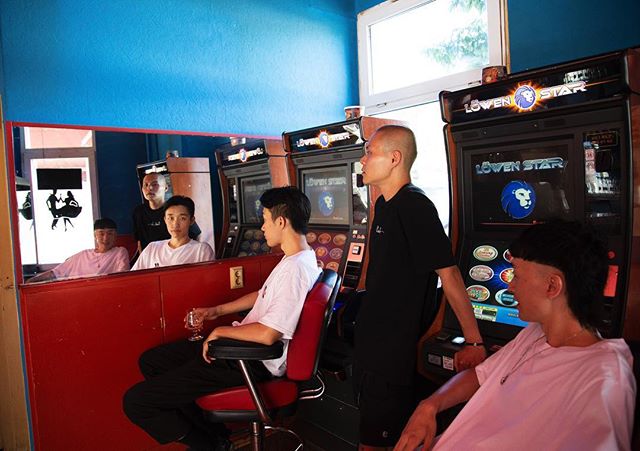 Four asian men are gathered around vintage arcade machines in a barbershop, reflected in a mirror.