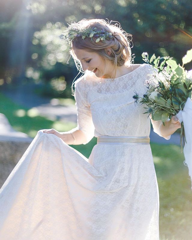 A smiling bride in a white lace wedding dress holds a bouquet in a sunlit garden.