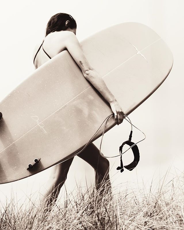 A woman walks through beach grass carrying a surfboard in a vintage-style monochrome image.