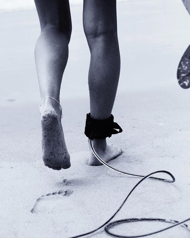A surfer walks on the beach, ankle leash on the right foot, with a board's edge visible.