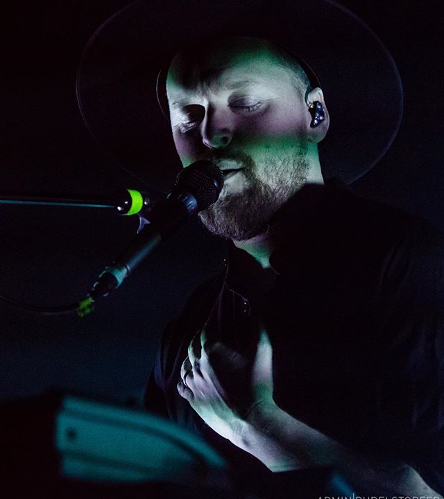 A man wearing a hat sings into a microphone on a dimly lit stage. 