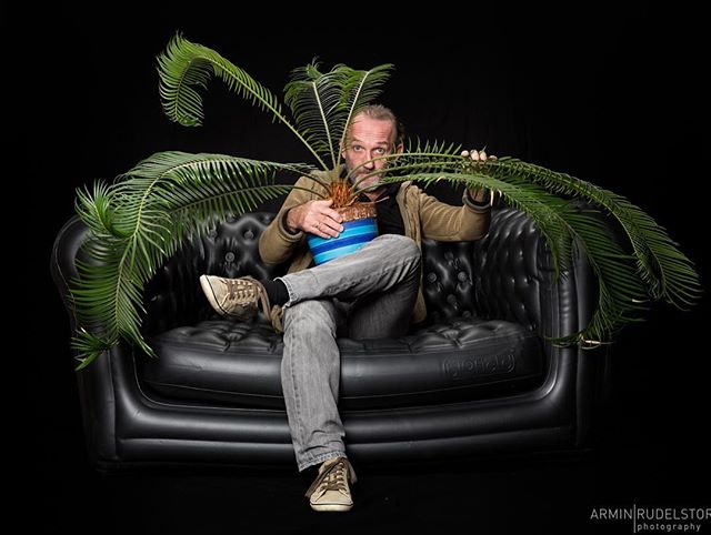A man sits on a black couch, holding a potted plant with large green fronds in a studio setting.