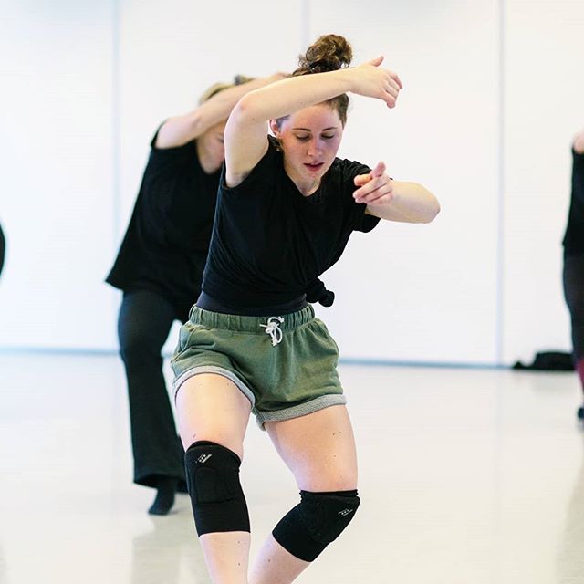 A woman dances in a studio wearing knee pads, shorts, and a t-shirt with other dancers in the background.
