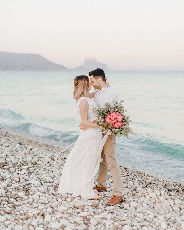 A bride and groom embrace on a pebble beach, the ocean behind them in this romantic wedding portrait.