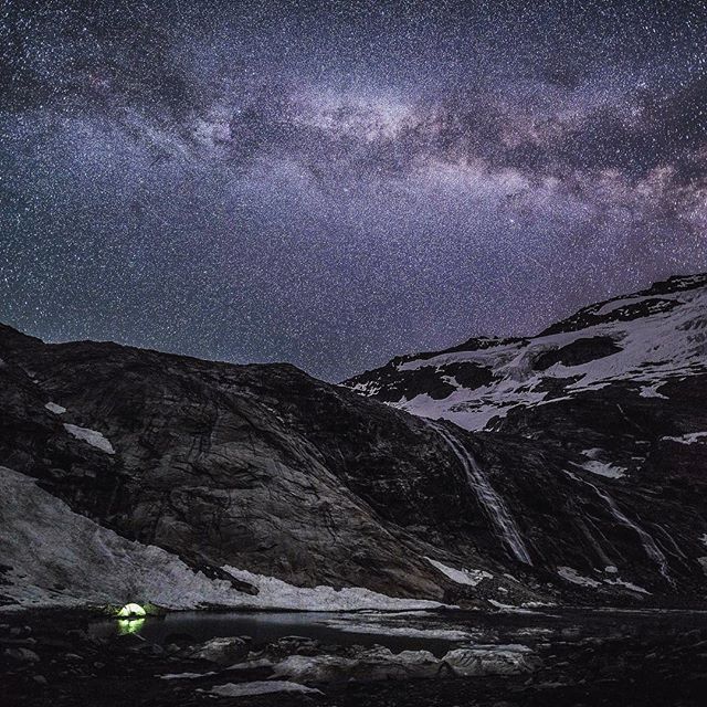 A tent glows at night next to a lake beneath a starry sky and mountains. A waterfall cascades down the slope.
