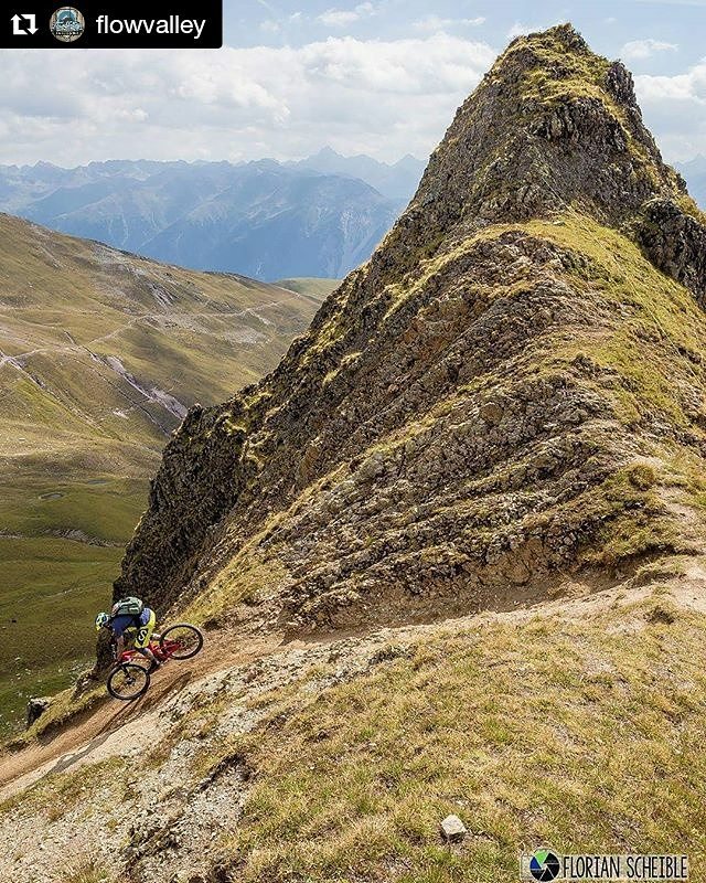 A mountain biker rides along a narrow trail on a steep, grassy mountain ridge with distant peaks.