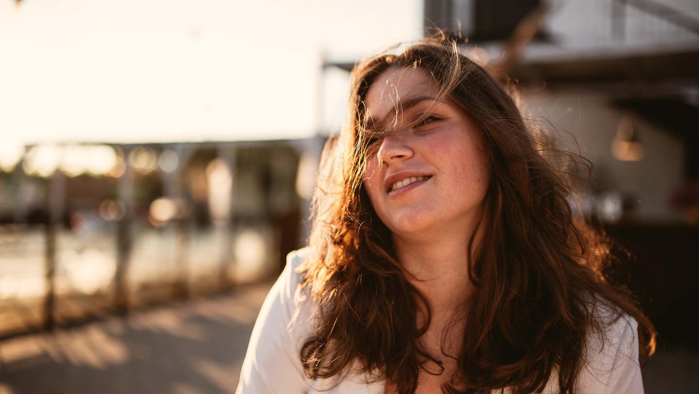 A smiling woman enjoys the sunlight in a natural, candid outdoor portrait with a soft, warm aesthetic.