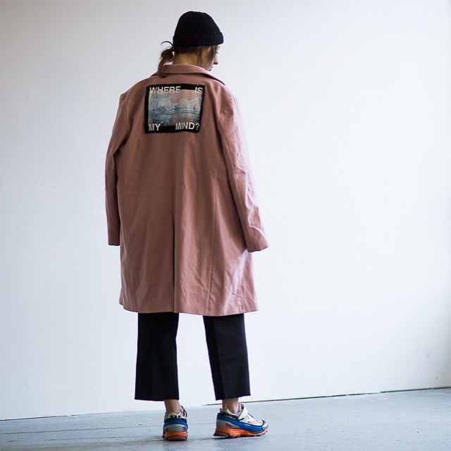 A woman stands in a studio wearing a pink coat with a graphic patch and black pants.